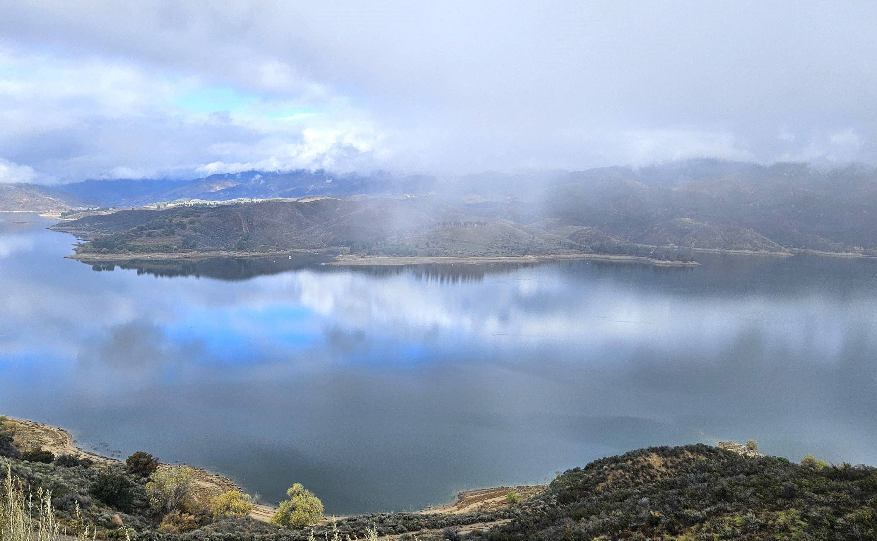 An image of a lake during scattered storms. More sky is visible in the reflection off of the lake than the in the sky.